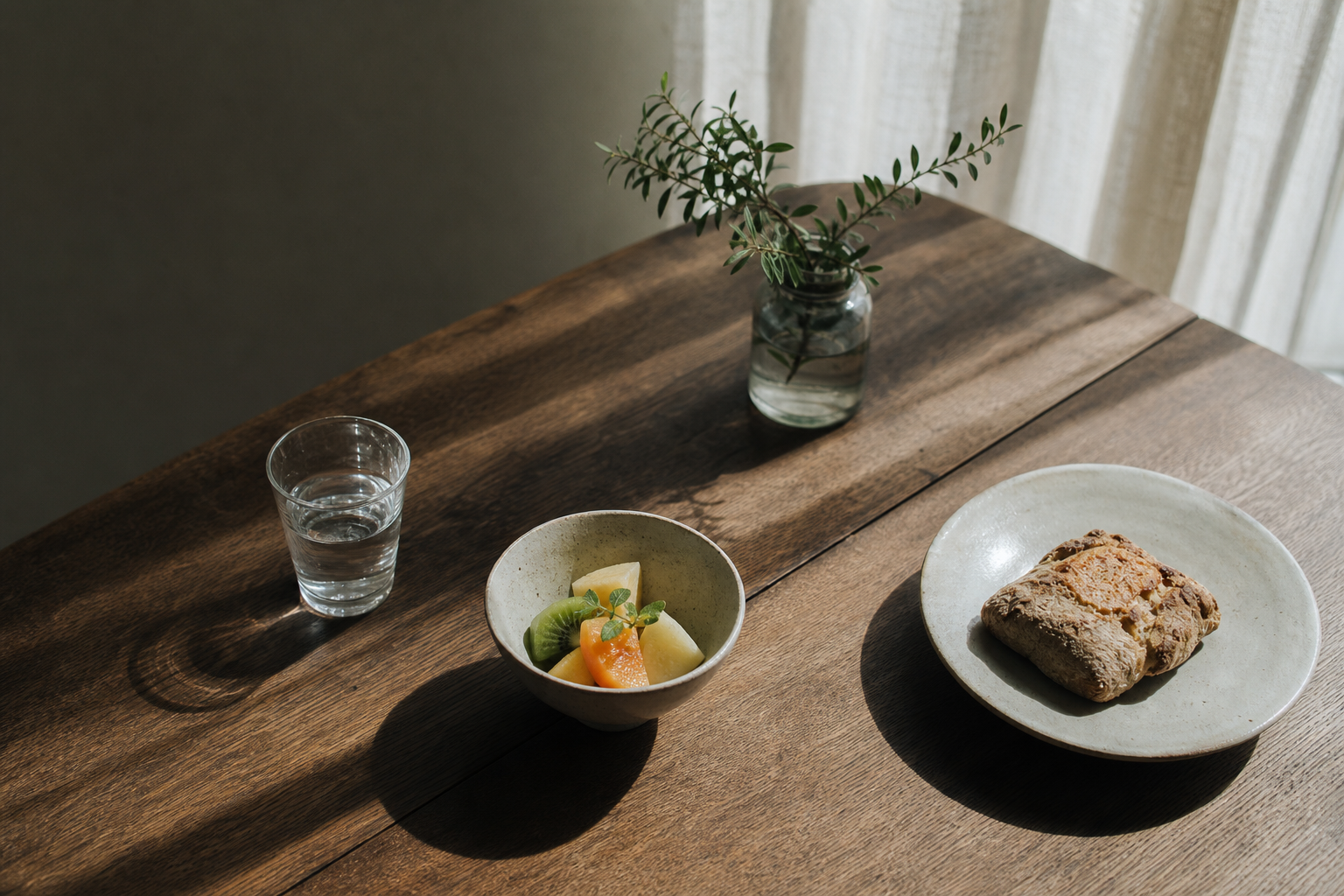 木の机に並んだ水のグラス、果物の小鉢、全粒粉のパン、一輪挿し。朝の光が差す静かな食卓